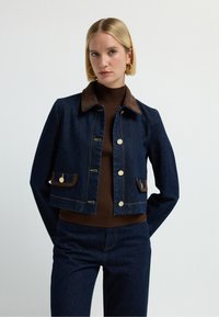 Džínová bunda s hnědým kordovým límcem, předními kapsami, zlatými knoflíky a kontrastním šitím. Nosená přes hnědý žebrovaný top.