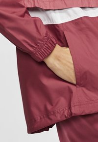 Veste de pluie bordeaux avec poignets élastiqués. Elle présente une bande blanche horizontale près des épaules et une poche latérale visible à la taille.