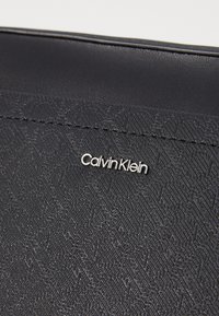 Must must handbag, mille kujundatud geomeetrilise mustriga, sileda tekstuuriga ja hõbedase "Calvin Klein" logoga detailiga.