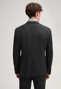 Dunkelgrau maßgeschneiderte Jacke mit glatter Textur, Reverskragen und einer einzelnen Rückenbelüftung. Kombiniert mit einem hellblauen Hemd mit Kragen darunter.