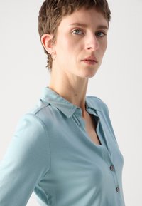Heledat sinine polo särk, mis on valmistatud siledast kangast, millel on nööbiga püstkrae, krae ja peened õmblusdetailid.