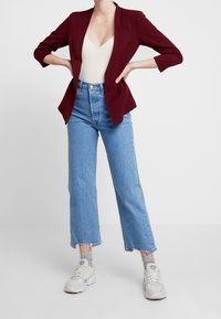 Blazer bordeaux con un design aderente, abbinato a un top lavorato crema e jeans a gamba dritta leggeri con vita alta. Sneakers bianche.