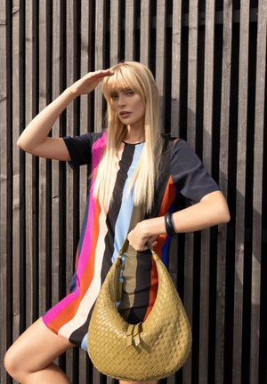 Woman in colorful striped dress holding beige woven handbag, shielding eyes from sunlight, standing against vertical wooden slatted wall.