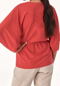 Haut ample corail avec manches larges et taille froncée. Texture douce, encolure arrondie et ourlet lisse. Associé à un pantalon beige.