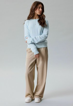 Jonge vrouw met lang bruin haar, gekleed in een lichtblauwe trui, beige broek met wijde pijpen en witte sneakers, staand tegen een effen grijze achtergrond.