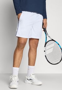 Biele športové šortky so bočnými vreckami, v kombinácii s bielymi teniskami a členkovými ponožkami. Drží čiernu a modrú tenisovú raketu.