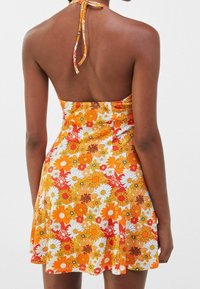 Nainen, jolla on ylisuurihana kaula-avoin mekko, jossa on oranssi, keltainen ja valkoinen kukkakuosi, nähtynä takaa.