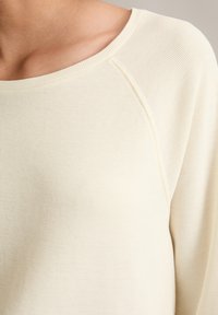 Light beige textured sweater with a wide neckline and raglan sleeves. The fabric appears soft and is finely ribbed.