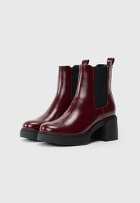 Botas de tornozelo em couro bordô com laterais elásticas pretas, bico arredondado e um salto grosso preto. Textura suave e design minimalista.