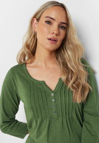 Maglietta verde a maniche lunghe con scollo a V, pattina con bottoni e pieghe verticali. Texture morbida con finitura liscia. Vestibilità casual.