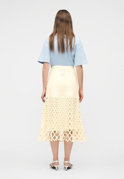 Femme aux cheveux bruns raides portant un haut bleu clair à manches courtes, une jupe midi en crochet crème et des sandales blanches à talons, debout de dos.