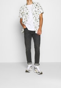 Kortärmad vit skjorta med gröna växtmönster, ihop med mörkgrå slim-fit jeans och vita sneakers med svarta och gula detaljer.