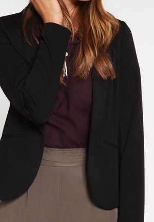 Femme portant un blazer noir sur un haut violet foncé et un pantalon beige taille haute, la main touchant ses cheveux.