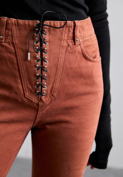 Roodbruine jeans met een hoge taille, voorzien van een vetersluiting aan de voorkant met zwarte koorden en metalen accenten. Textuurstof met vijf zakken.