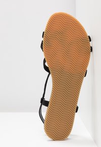 Semelle de sandale avec bande de roulement en caoutchouc beige présentant un motif en zigzag, sangles noires visibles sur les côtés, sur fond blanc.