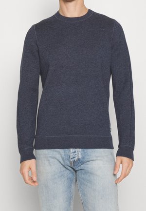 Mann trägt einen dunkelblauen Pullover mit Rundhalsausschnitt und langen Ärmeln sowie hellblaue ausgewaschene Jeans, steht vor einem schlichten hellen Hintergrund.