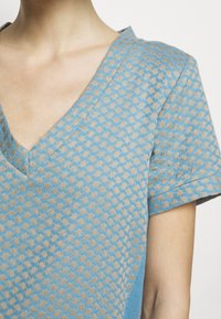 Modrý top s výstřihem do V a krátkými rukávy, s texturovaným vzorem béžových tvarů, který zdůrazňuje kontrast mezi barvami a materiály.