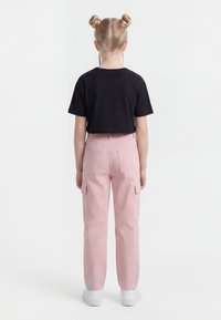 Két fonott kontyot viselő gyermek áll háttal, fekete rövid felsőt, rózsaszín terápiás nadrágot és fehér sportcipőt visel fehér háttér előtt.