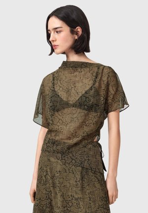 Jonge vrouw met kort donker haar, gekleed in een doorschijnende olijfgroene blouse met patroon over een zwarte beha en bijpassende rok, kijkend naar links.