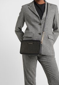Persona che indossa un completo a pesce d'oro grigio e una camicia nera, mentre porta una borsa a tracolla nera con borchie e tracolla dorata.