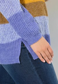 Ręka spoczywa na kieszeni niebieskich dżinsów, na sobie ma sweter z prążkowanej dzianiny w kolorze lawendowym, białym i musztardowym, z bocznym rozcięciem.