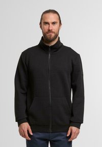 Mann mit zurückgebundenem Haar und Bart, der eine schwarze strukturierte Zip-Up-Jacke und dunkle Hosen vor einem schlichten hellen Hintergrund trägt.
