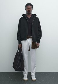 Haut à capuche noir zippé, polo foncé à côtes, pantalon blanc, sac en bandoulière marron, baskets blanches avec accents beiges, debout sur une surface grise.