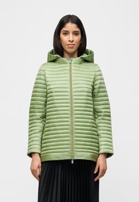 Γυναίκα με ανοιχτοπράσινο καπιτονέ μπουφάν με κουκούλα και φερμουάρ, συνδυασμένο με μαύρη πλισέ φούστα, σε απλό ανοιχτό φόντο.