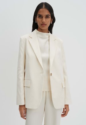 Femme aux longs cheveux foncés portant un blazer crème sur un haut à col haut et un pantalon assorti, sur un fond clair uni.