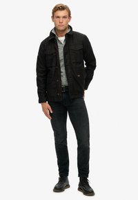 Sort yderjakke med lommer, lagd over en ternet skjorte. Mørke jeans og snøreløse sko fuldender outfittet. Enkel design, holdbart stof.