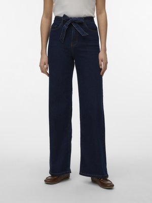 Donna che indossa jeans blu scuro a vita alta e gambe ampie con cintura annodata, top bianco e scarpe di pelle marrone, in piedi davanti a uno sfondo semplice.