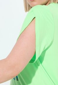 Neon grøn kortærmet top med en afslappet pasform. Glat tekstur og buede ærmedetaljer. Har et diskret design på forsiden.