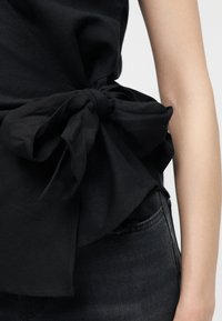 Μαύρο ύφασμα δεμένο σε μεγάλο φιόγκο στη μέση μιας μαύρης μπλούζας που φοριέται με μαύρο τζιν, με ορατό χέρι στα δεξιά.