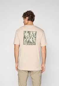 Beige bomull t-skjorte med et rektangulært grønt camo-graphic med "Columbia"-logoen på ryggen. Korte ermer og rund halsdesign.