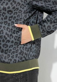 Leopardmönstrad bomberjacka med en mörkgrå satinfinish, med en grön ribbad nederkant och gula detaljer vid ärmslutet. Sidoficka synlig.