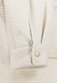 White fuzzy fabric with a textured finish. Cuffs feature a gold button detail, adding a subtle accent to the design.