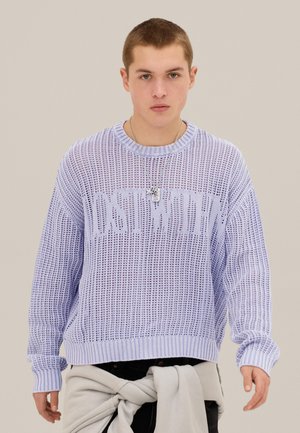 Mladý muž v svetlofialovom pletenom svetri s nápisom "LOST TOKYO", so svetlosivou bundou zviazanou okolo pása a strieborným náhrdelníkom.