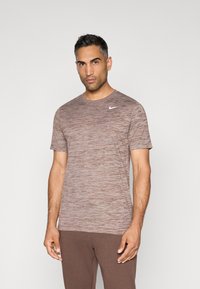 Camiseta deportiva marrón jaspeada con mangas cortas y cuello redondo; presenta un pequeño logo blanco de Nike en la parte izquierda del pecho.