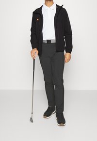 Chaqueta negra con cremallera, camisa polo blanca, pantalones grises y zapatos de golf negros. Sosteniendo un palo de golf, de pie sobre un fondo blanco.