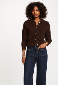 Brauner, strukturierter, knöpfbarer Cardigan mit Kragen, versehen mit silberfarbigen Knöpfen, getragen über dunkelblauen Jeans mit Akzentnähten.