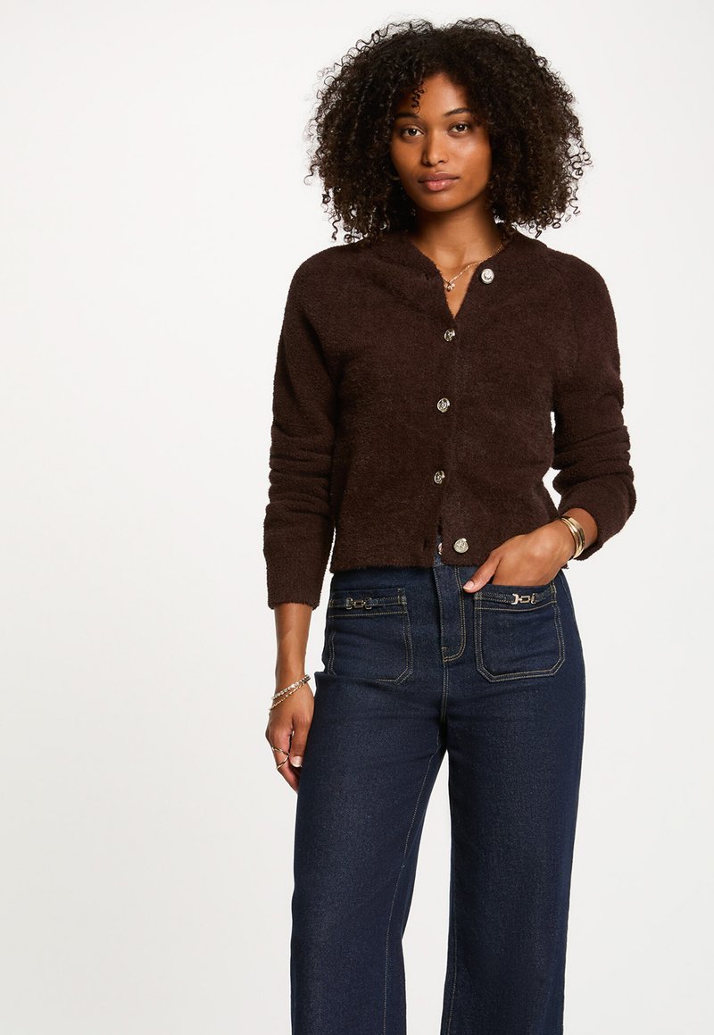 Brauner, strukturierter, knöpfbarer Cardigan mit Kragen, versehen mit silberfarbigen Knöpfen, getragen über dunkelblauen Jeans mit Akzentnähten.