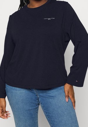 Femme portant une chemise Tommy Hilfiger bleu marine à manches longues avec logo sur la poitrine et le poignet, associée à un jean bleu clair.