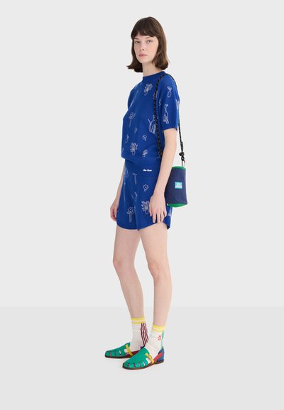 Kobieta ubrana w niebieską, krótką bluzkę z nadrukiem i szorty, niosąca granatowo-zieloną torebkę na ramię, z kolorowymi skarpetkami i zielonymi sandałami.