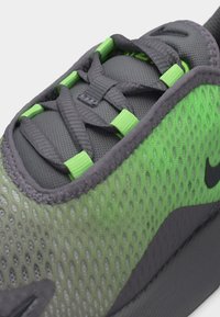 Κοντινή λήψη γκρι και πράσινου αθλητικού παπουτσιού με δίχτυ, με μαύρα κορδόνια που περνούν μέσα από πράσινα πλαστικά κρίκους και ορατό το μαύρο λογότυπο Nike.
