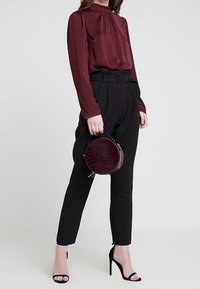 Burgundy långärmad blus med samlad halsringning, svarta högmidjade byxor och en rund, texturerad mörkröd handväska. Svarta högklackade skor.
