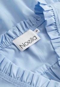 Jasnoniebieska tkanina z falbanami; posiada białą metkę z napisem "Noella" oraz rozmiarem "S". Gładka tekstura, minimalne detale.