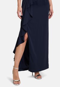 Mörkblå, golvlång klänning med volangdetaljer och en slits på sidan, parat med silverfärgade högklackade sandaler med ankelsnodd.