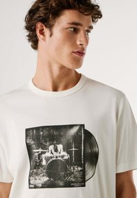 Witte katoenen t-shirt met een zwart-witprint van een drummer en een vinylplaat. Ronde halslijn en korte mouwen.