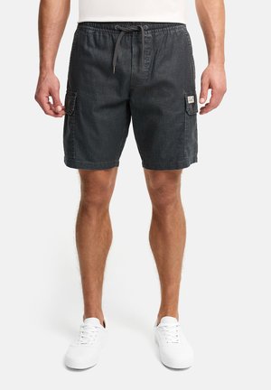 Mann iført svarte cargoshorts med snor i midjen og hvite joggesko, stående mot en ensfarget bakgrunn.