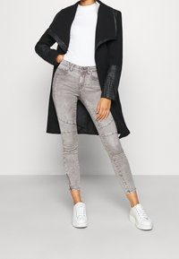 Abrigo negro con mangas de cuero, top blanco acanalado, jeans ajustados grises con cremalleras en los tobillos y zapatillas blancas.